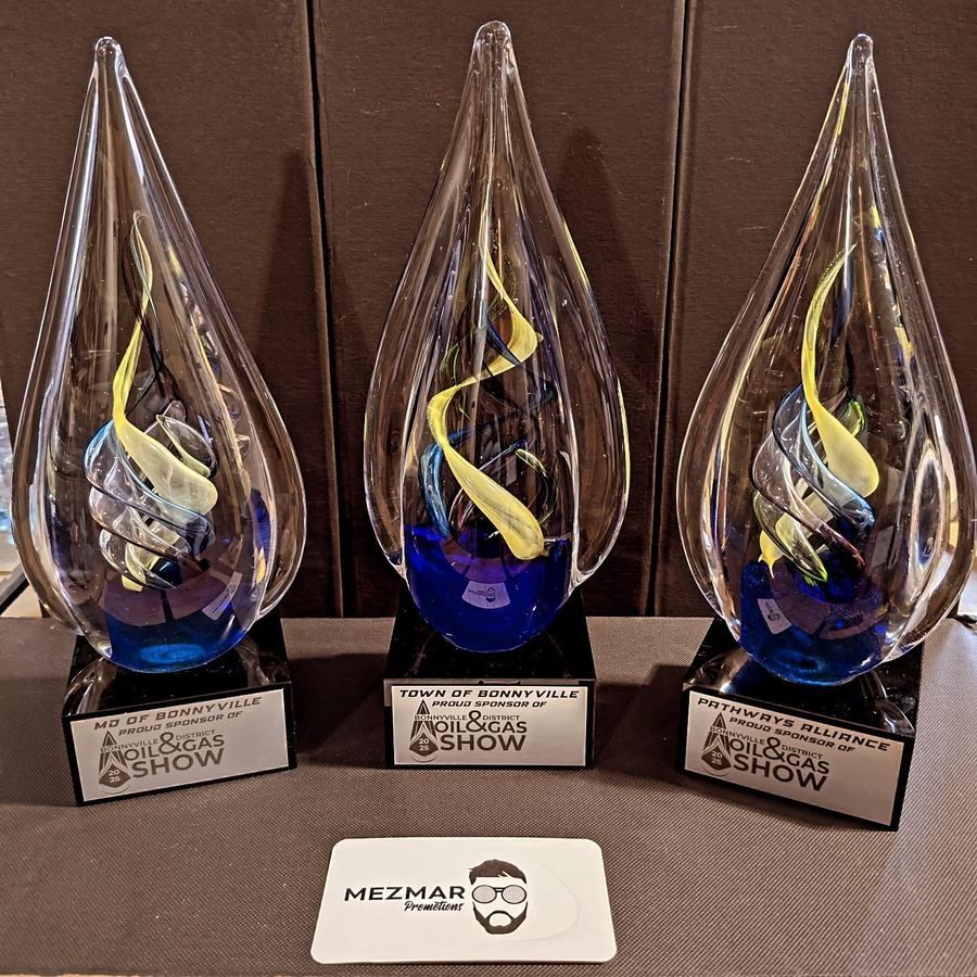 Three hand-blown glass teardrop awards on black bases with engraved plates, beside a Mezmar Promotions business card